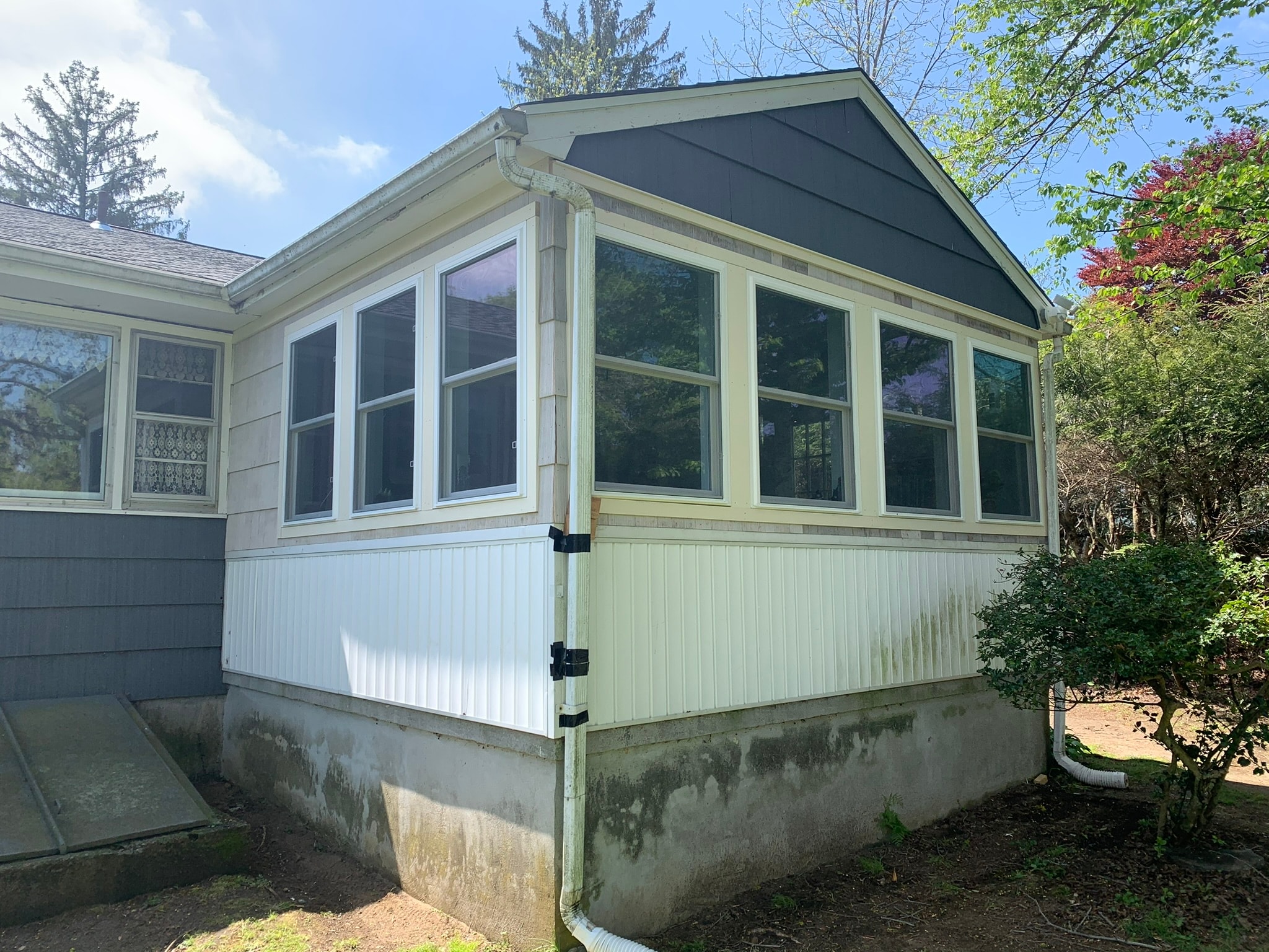 Corner view of completed four-season porch conversion — new double-hung windows wrapping front and side walls, cedar shake siding, dark gable end, concrete foundation, spring foliage Eastern Connecticut