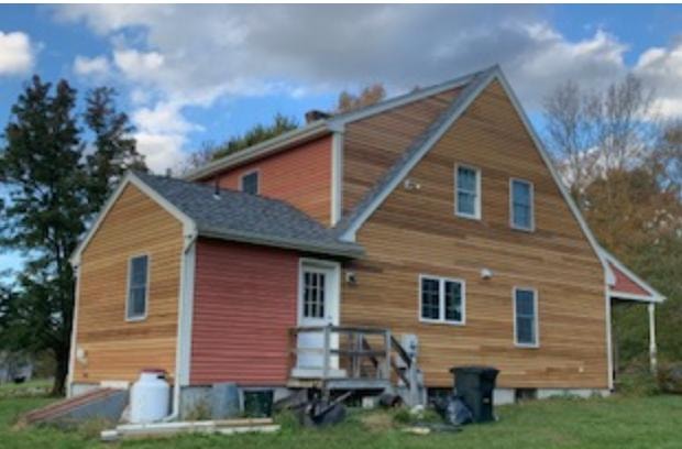 Wide view of new clear grade cedar siding installed on south side of farmhouse — two-story gable end and lower addition fully re-sided, original red-painted sections visible on adjacent walls, Eastern Connecticut