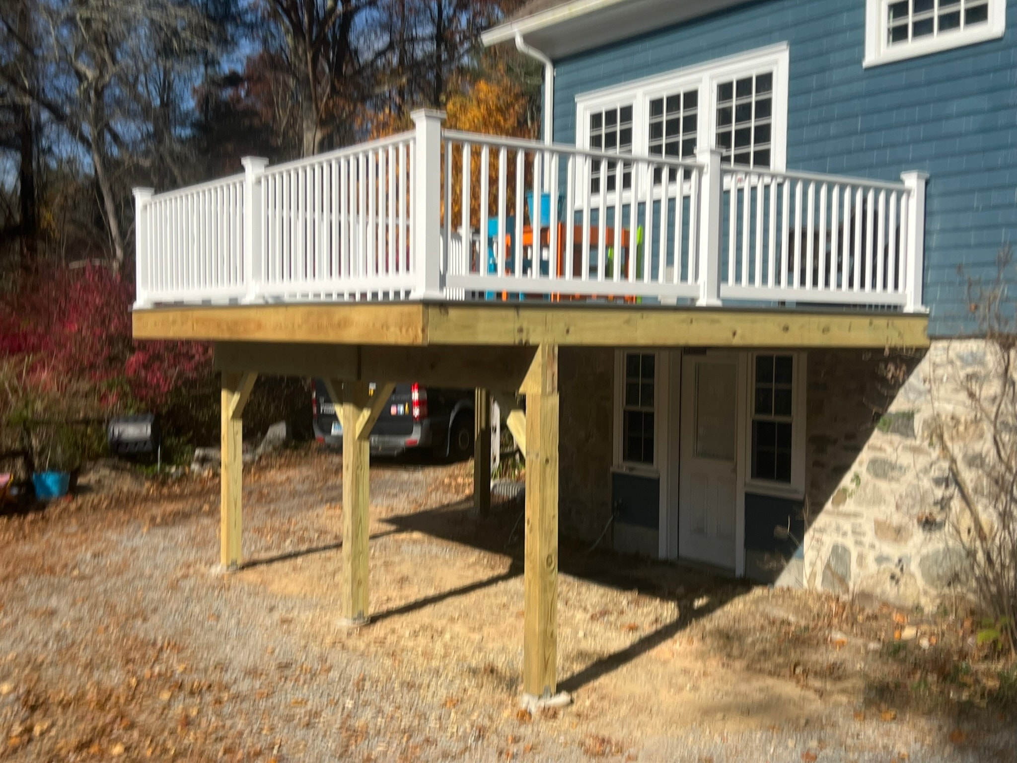 Elevated deck framing complete — pressure-treated posts and joists, white composite railing installed, new entry door cut into small porch below deck, blue cedar-shake colonial home Eastern Connecticut