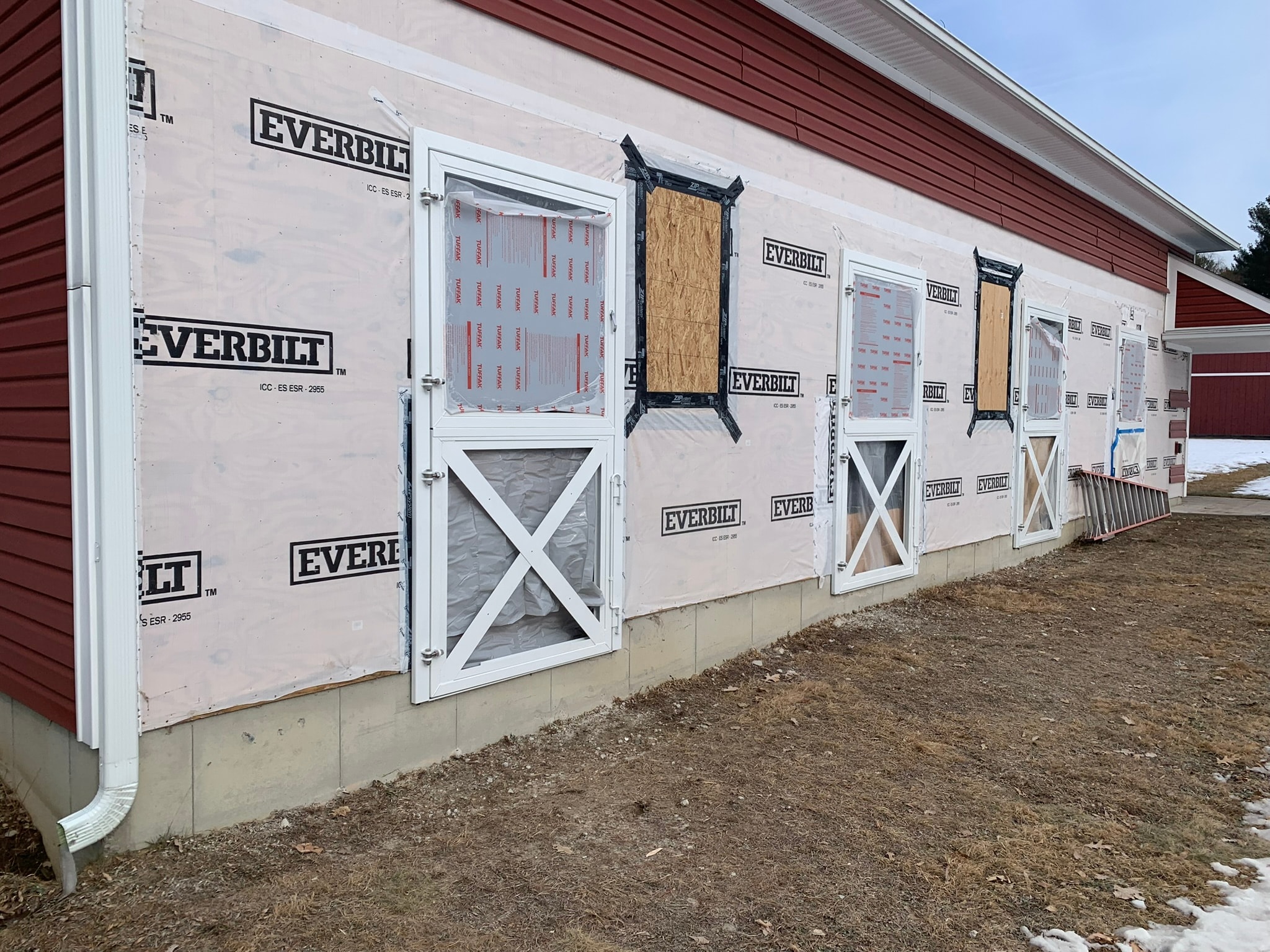 Horse barn exterior during stall door installation — Everbilt house wrap on red siding, white X-brace stall door frames being set, construction phase, Eastern Connecticut