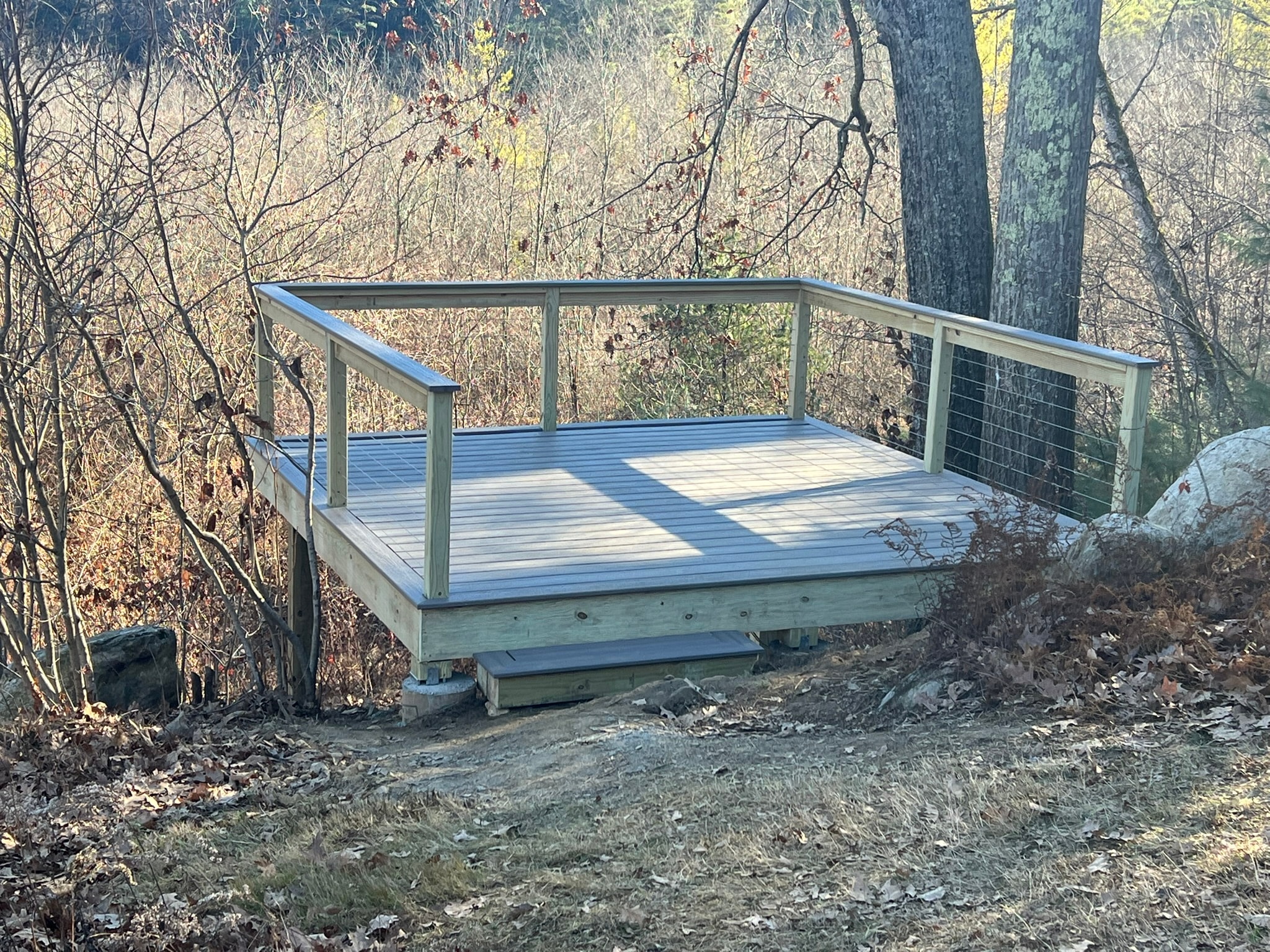 Completed pond overlook deck built into steep rocky embankment — gray PVC decking, PT framing on concrete footings, wire panel railings on all sides, fall foliage Eastern Connecticut