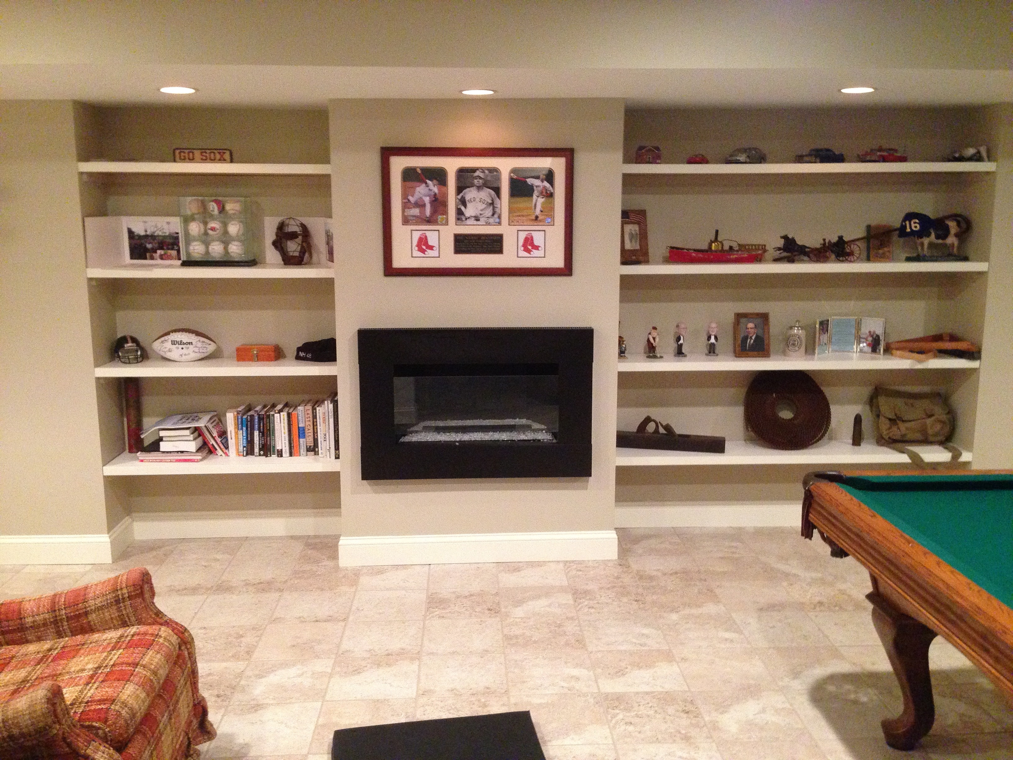 Finished basement entertainment wall — custom built-in shelving flanking center TV niche, display collectibles and books, recessed lighting, tile ceiling panels, antique pool table at left, Eastern Connecticut