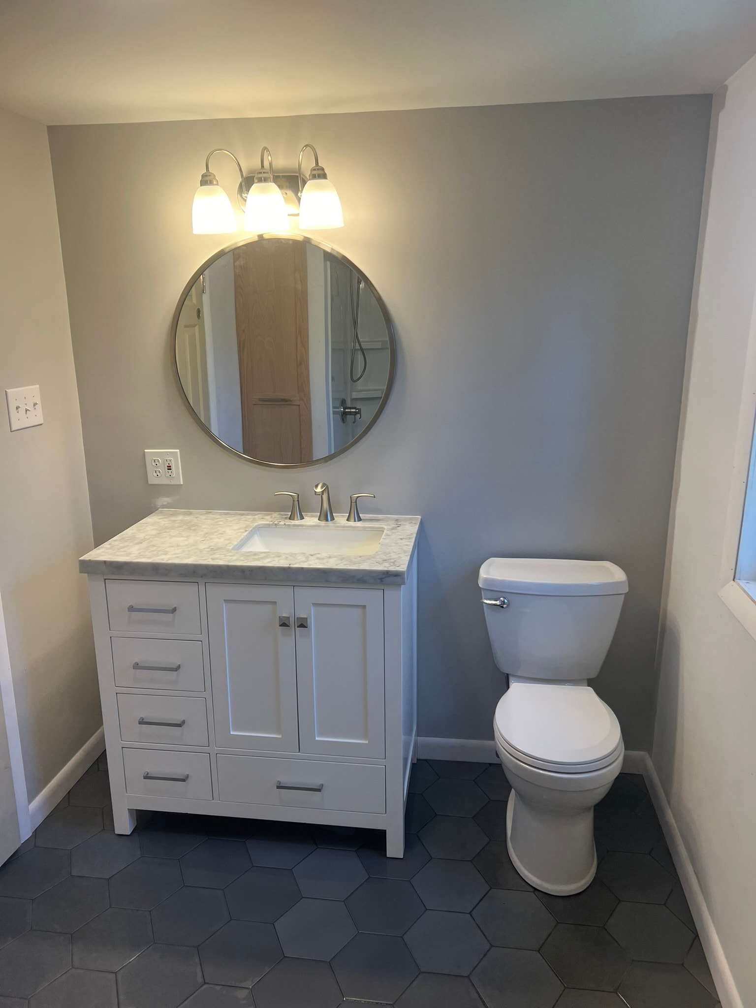 Finished full bathroom — new white shaker vanity with marble countertop, round mirror, and three-light fixture over black hexagon tile floor