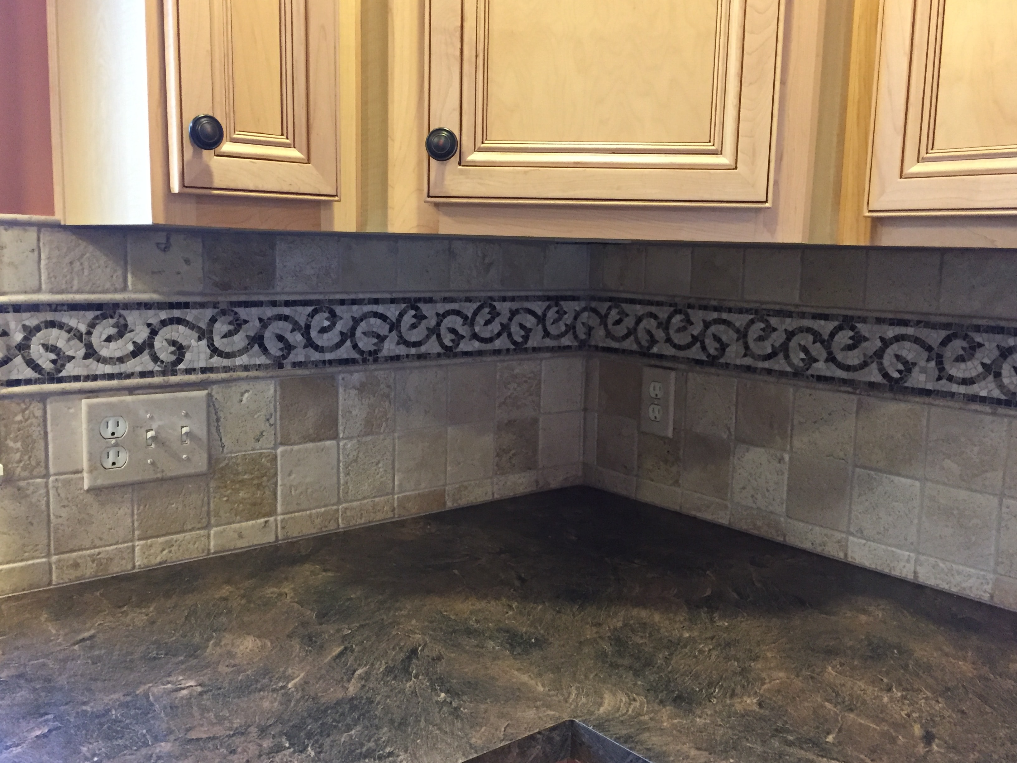 Kitchen backsplash corner detail — tumbled travertine tile with Italian mosaic scroll border and dark granite countertop