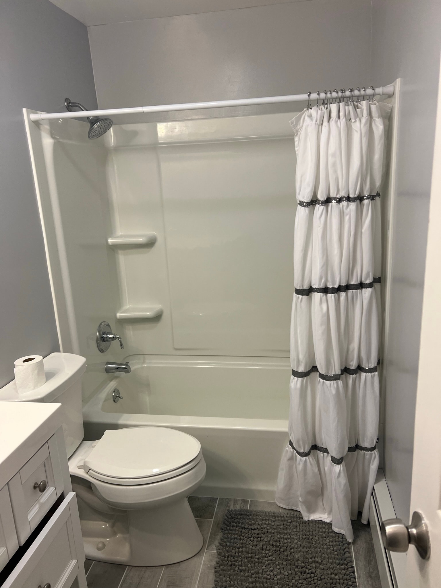 Finished small bathroom gut remodel — new tub/shower unit with chrome fixtures and ruffled shower curtain, gray wood-look tile floor