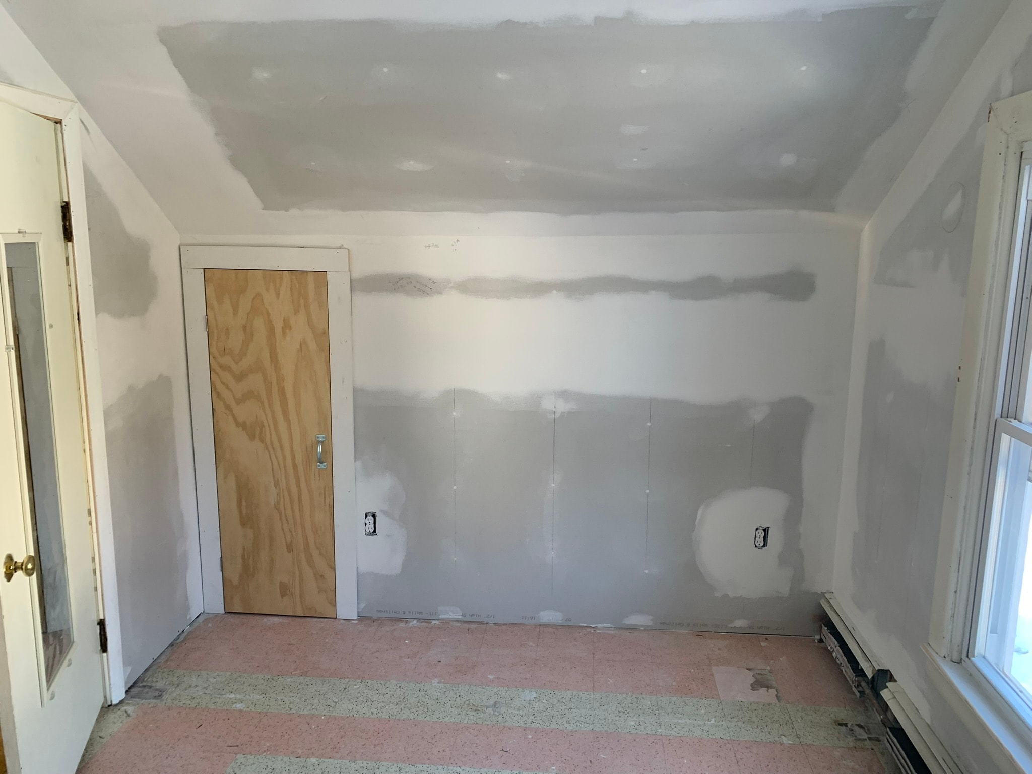Victorian second-floor dormer bedroom after drywall and compound — sloped ceiling with double-hung windows, new baseboard trim, original teal and tan checkerboard tile floor, radiator, new electrical outlets, Eastern Connecticut